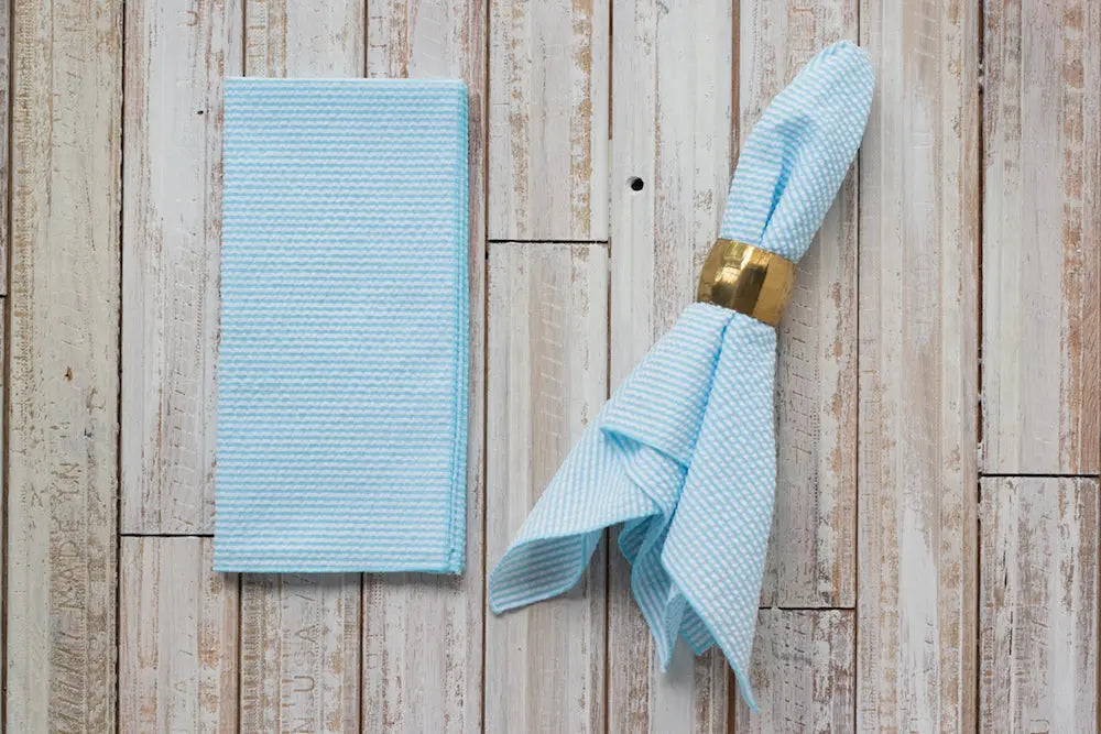 Light blue seersucker napkins, one folded, one with gold ring, on wood.