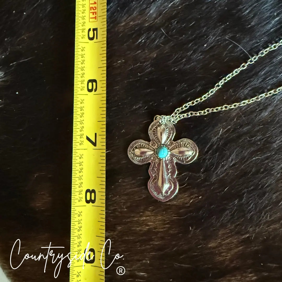 Silver Southwestern cross necklace with turquoise stone next to measuring tape.