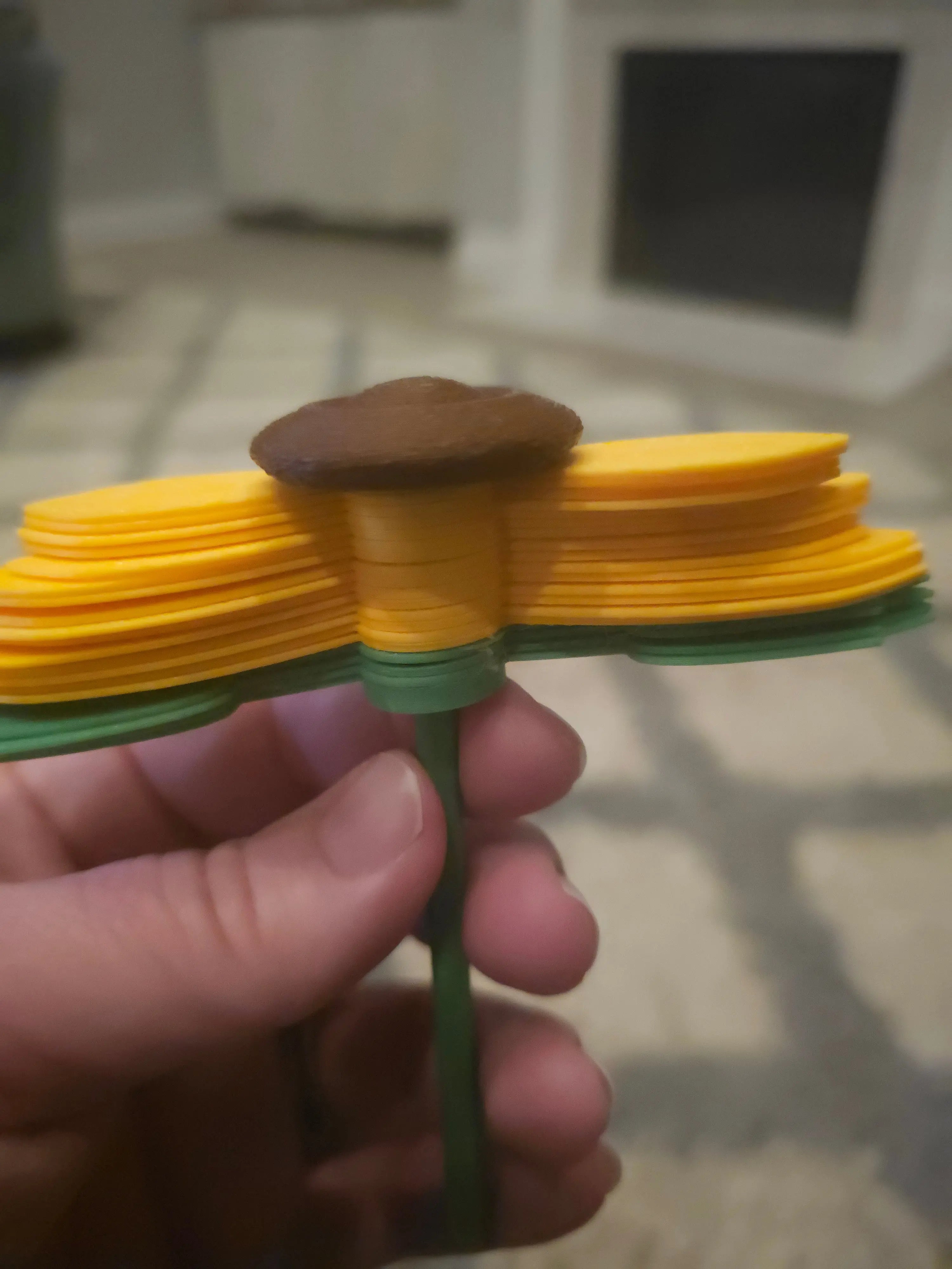 Hand holding a 3D printed sunflower fidget spinner with brown center, yellow petals, and green stem. Sensory toy for kids.