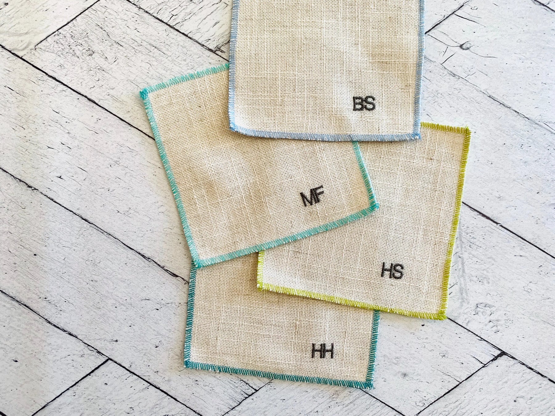 Linen cocktail coasters with monograms BS, MF, HS, and HH on a wooden background.
