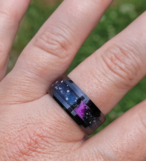 Hand wearing a black ceramic ring with embedded ashes, onyx mica powder, glowing star constellations, and purple glow.