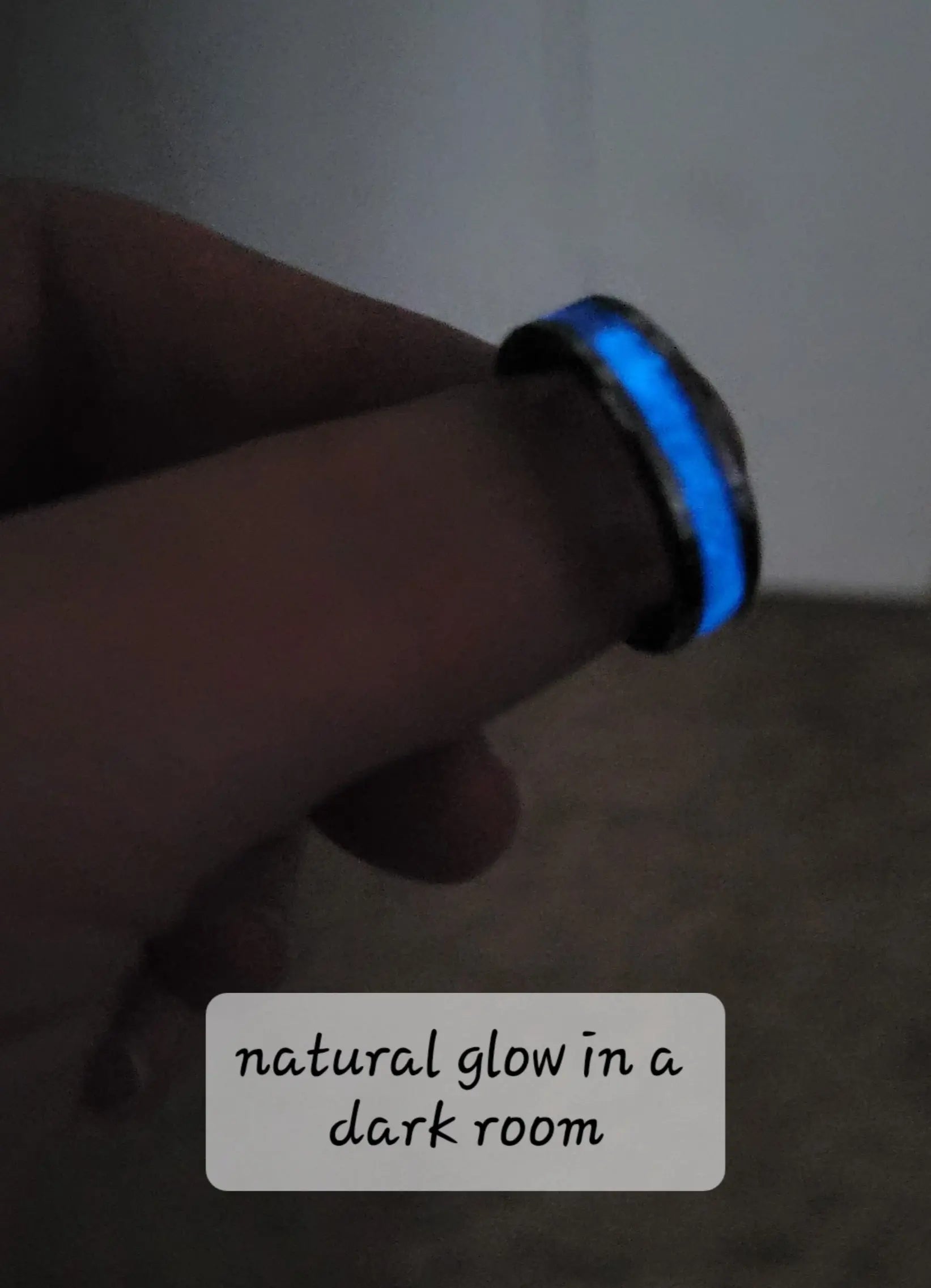 Glowing blue memorial ring with ashes on a finger in a dark room, viewed at an angle to see the band.