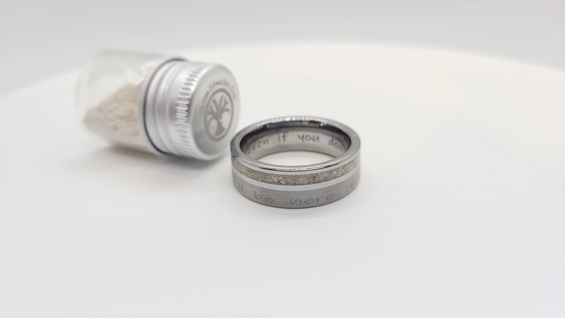 Tungsten memorial ring with ashes inlay and inscription \"Even if you don't, I'll be here.\" next to ashes in glass container.