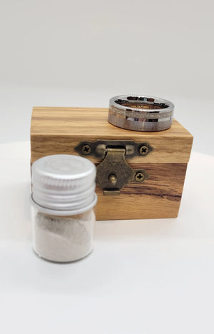 Tungsten memorial ring with ashes on wood box with small jar on white backdrop.