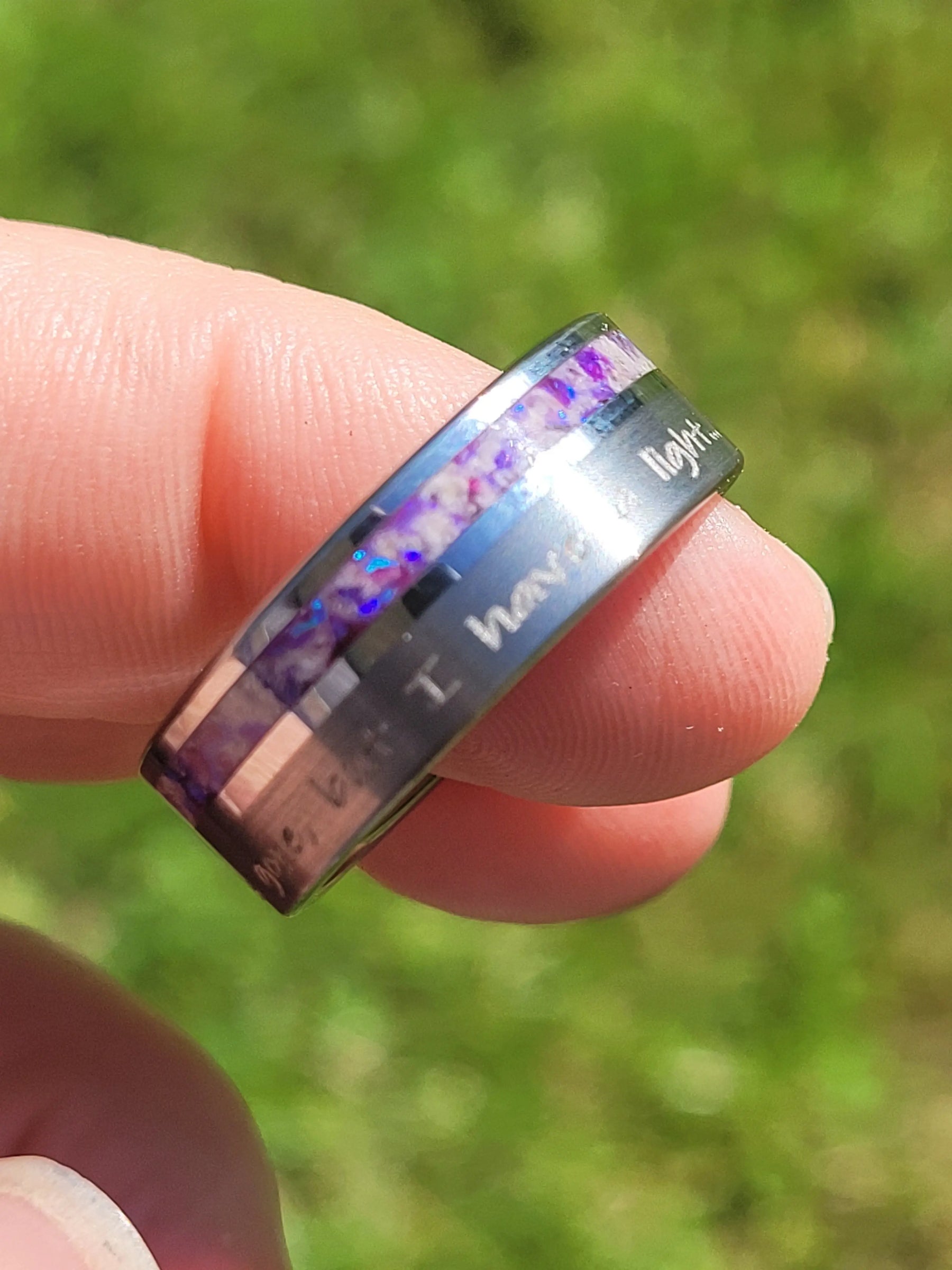 Memorial ring with purple opal inlay held in fingers. Engraved tungsten band contains ashes.