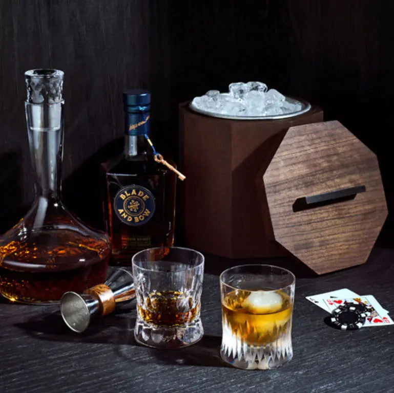 Wooden ice bucket with lid, whiskey decanter and bottle, two glasses of liquor, playing cards, and ice. 