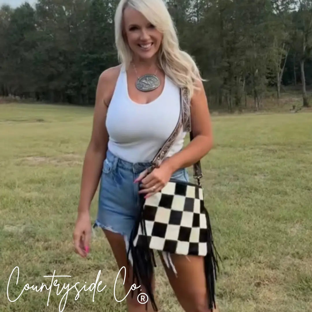 Woman in denim shorts and white tank top with a checkerboard fringe bag and NFR buckle necklace.