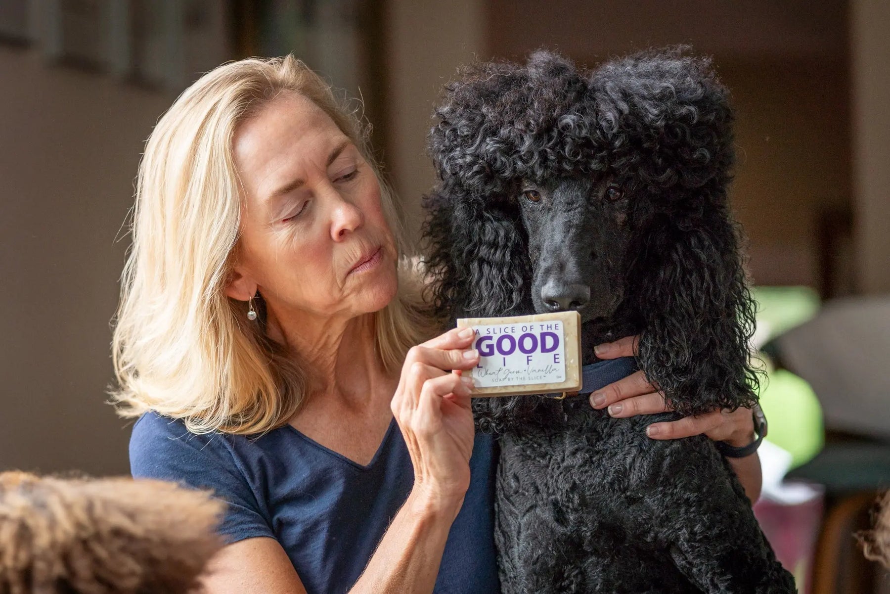 Woman holding a black poodle and a bar of wheat germ and vanilla soap.