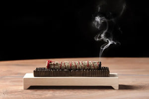 Wooden incense holder with bronze casting holding sage bundle, smoke rising against black background.