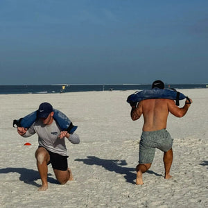 WORKOUT BATTLE BAG - SPARK STARTER: Two men training on a beach with sandbags.