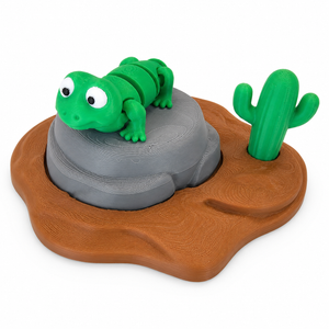 Green 3D printed lizard toy on a gray rock with cactus and brown desert base by The Tween Space