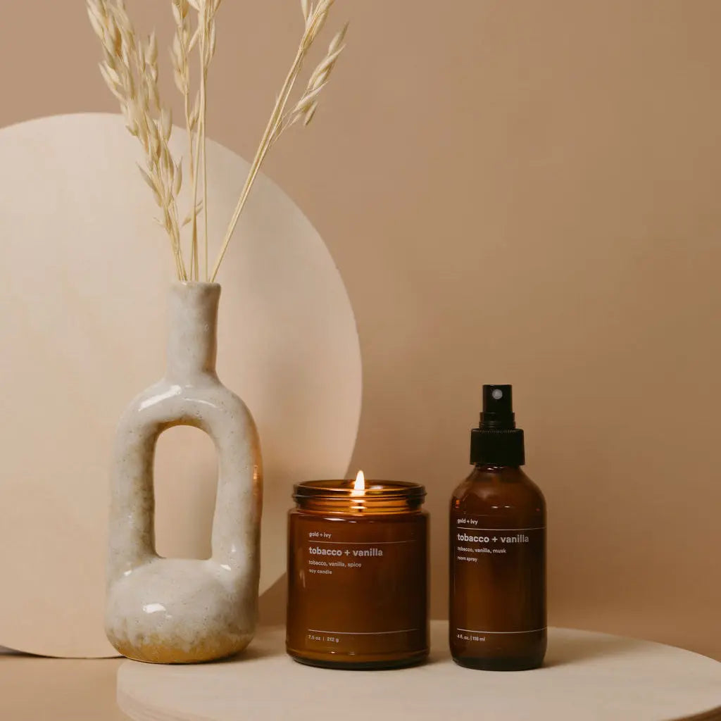 Lit Tobacco + Vanilla candle and room spray bottle next to a ceramic vase with dried grass.